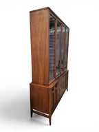 Mid Century Modern Broyhill Brasilia walnut breakfront China cabinet in a right-front view. Features the Brasilia collection's catseye sweeping arches and tapered legs, with brass hardware and glass display doors. Vintage as-is, gently refreshed, including credenza base with three drawers and two doors and top display deck with two doors.